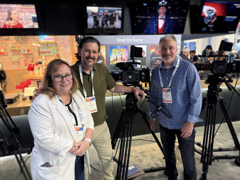 Three professionals standing at a Sony booth at NAB Show, posing with a Sony cinema camera mounted on a tripod, with “Shot on Sony” signage and production displays in the background.