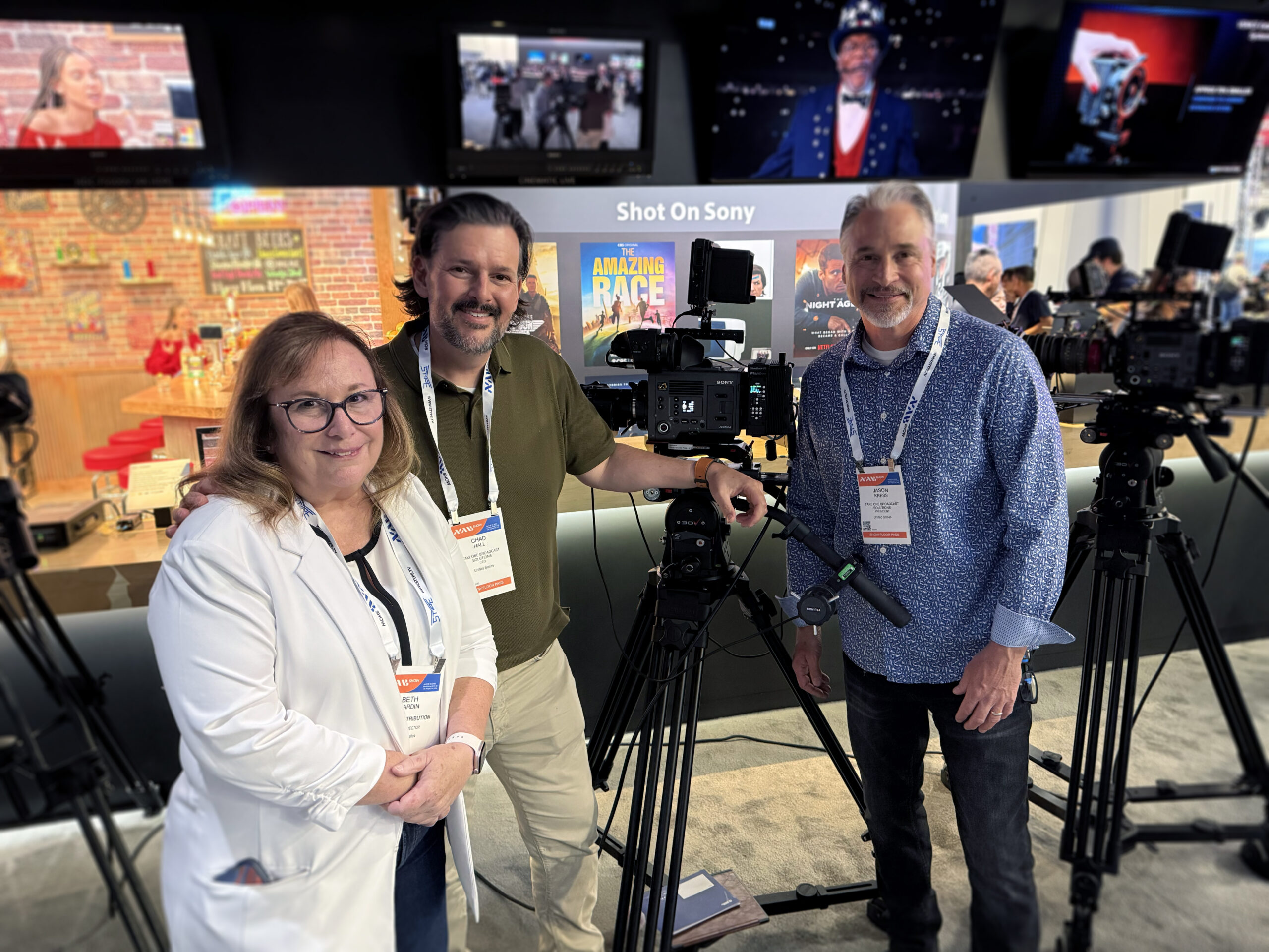 Three professionals standing at a Sony booth at NAB Show, posing with a Sony cinema camera mounted on a tripod, with “Shot on Sony” signage and production displays in the background.